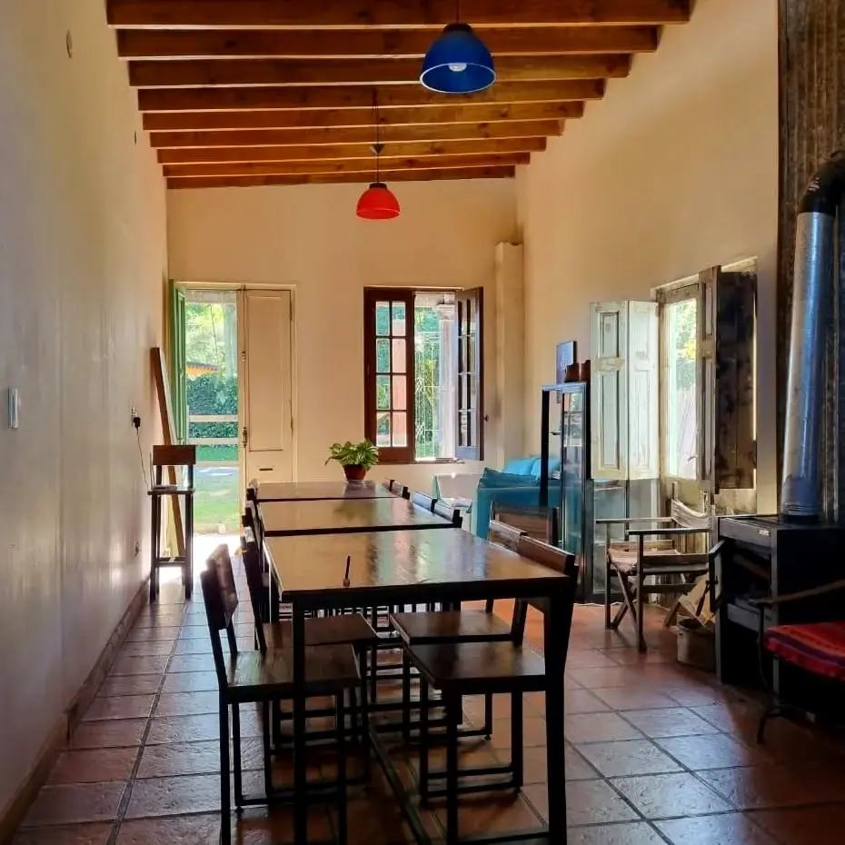 El comedor de la posada: mesas largas de madera, piso de baldosa terracota, techo de vigas y un sillón celeste junto a la ventana.