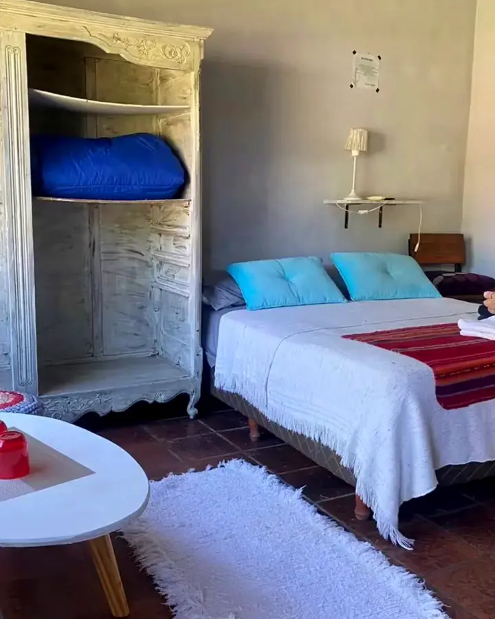 Una de las habitaciones: cama de dos plazas con almohadones celestes, un ropero blanco antiguo y piso de baldosa.