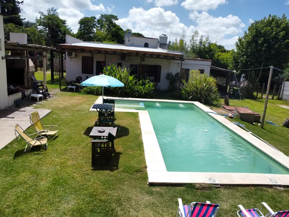 El parque de la posada en un día de sol: la pileta larga con reposeras, la sombrilla celeste y la casa blanca al fondo, entre árboles.